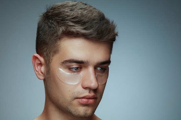 Close-up profile portrait of young man isolated on grey studio background. Male face with collagen patches under eyes. Concept of men's health and beauty, cosmetology, body and skin care. Anti-aging.