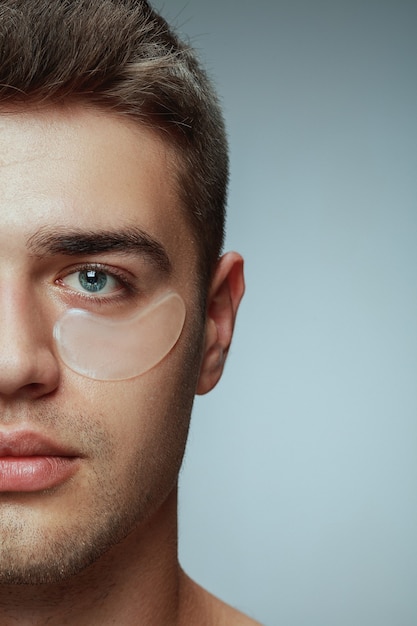 Close-up profile portrait of young man isolated on grey studio background. Male face with collagen patches under eyes. Concept of men's health and beauty, cosmetology, body and skin care. Anti-aging.