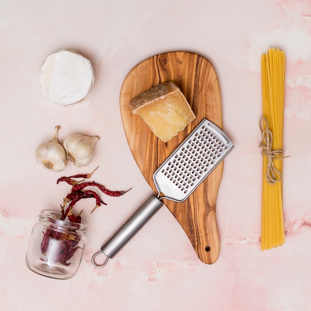 Free photo close-up of raw pasta; cheese; dried chili; garlic and kitchen utensil on pink backdrop