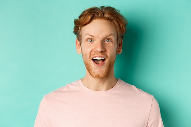 Free photo close up of redhead bearded man raising eyebrows and looking surprised at camera, checking out promotion, standing over mint background.