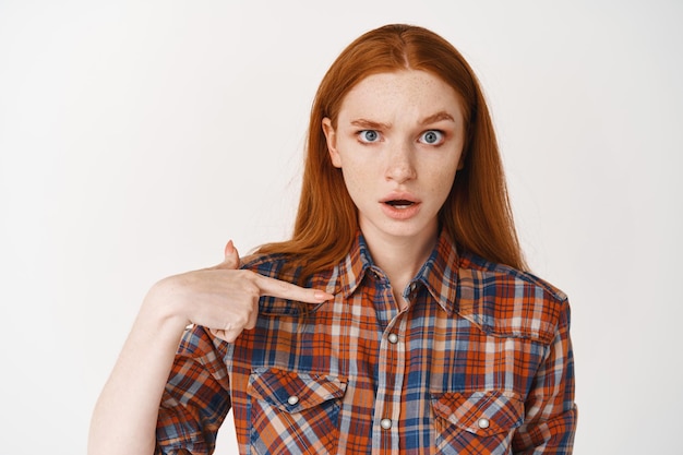 Close-up of redhead girl with pale skin looking confused, pointing at herself puzzled, standing on white wall