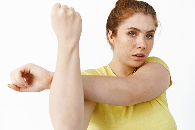 Крупный план рыжеволосой женщины больших размеров, растягивающей руки, разминающейся перед фитнес-тренировкой, мотивированной на камеру, стоящую на белом фоне