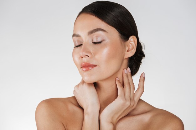 Close up of relaxed brunette woman with soft skin posing at camera with closed eyes, isolated over white