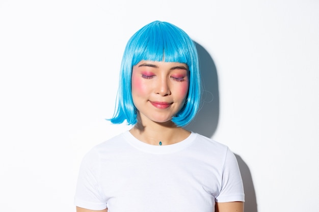 Close-up of romantic, cute asian woman in blue wig, close eyes and daydreaming with pleased smile, standing.