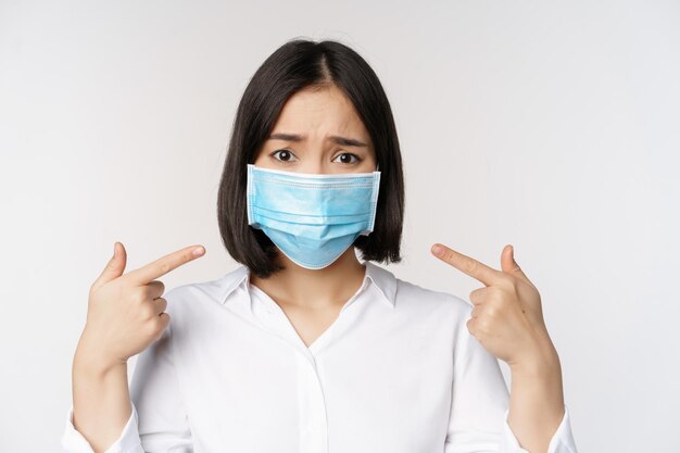 Close up of sad asian girl in medical face mask pointing at her head and looking upset standing over white background