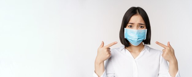 Close up of sad asian girl in medical face mask pointing at her head and looking upset standing over white background