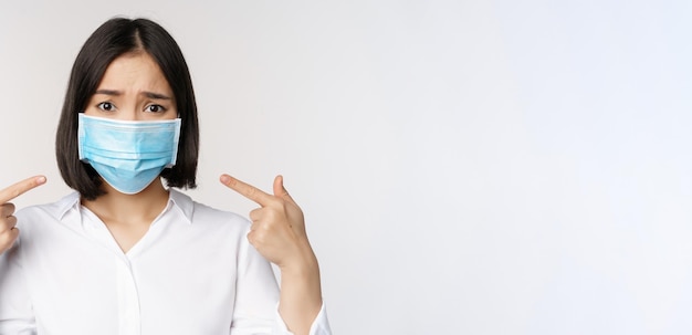 Крупный план грустной азиатской девушки в медицинской маске, указывающей на голову и выглядящей расстроенной, стоящей на белом фоне