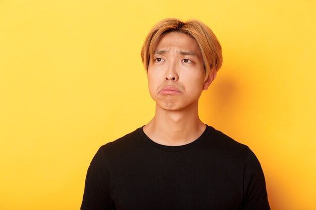 Close-up of sad and disappointed handsome asian guy sulking upset, looking upper left corner with regret or jealousy, standing yellow wall