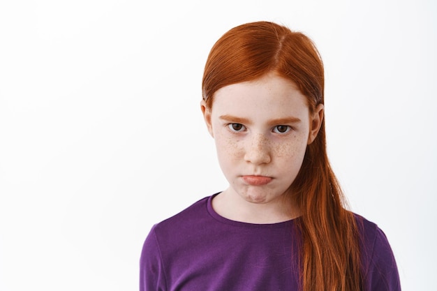 Free photo close up of sad redhead girl with freckles sulking, angry or gloomy looking at camera, being upset or jealous, frowning dipleased, standing over white background