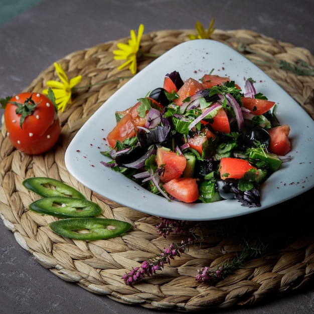 Салат крупным планом с помидорами, огурцами, листьями салата, луком, базиликом, оливками в белой тарелке на плетеной подставке