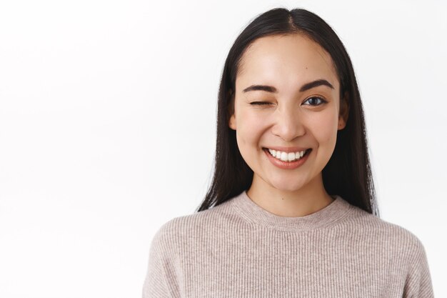 Close-up sassy, enthusiastic happy smiling asian woman with long dark hair, natural nude make-up, having great day, send positive vibes, winking to cheer friend up and say everything under control