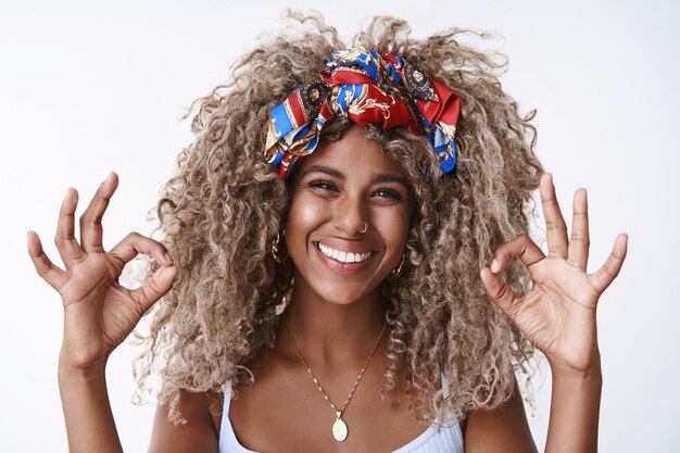 Close-up satisfied and happy, blond curly-haired girl with afro hairstyle, pierced nose, stylish headband, smiling approvingly, show okay, ok gesture pleased with good service, standing delighted