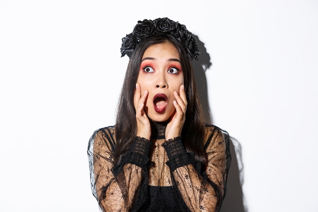 Free photo close-up of scared asian woman in halloween costume of witch looking frightened at upper left corner, gasping and standing ambushed over white background.