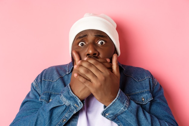 Close-up of scared Black man gasping, cover mouth and stare at camera afraid, standing over pink background.