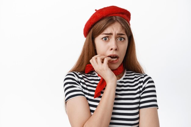 Close up of scared young woman in beret, biting fingernails and looking frightened and anxious at front, fear something, standing against white wall