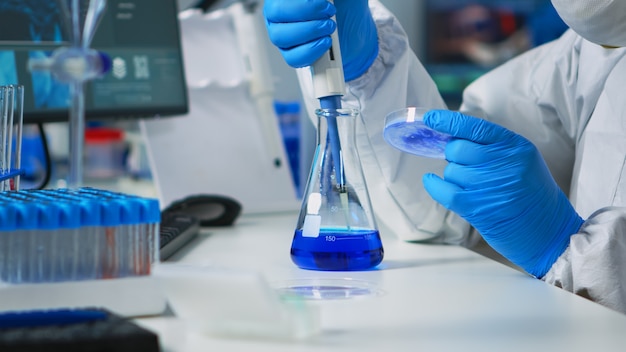 Close up of science technician in ppe suit using a pipette with a microtiter plate and a petri dish working in laboratory. Team examining virus evolution for vaccine development against covid19