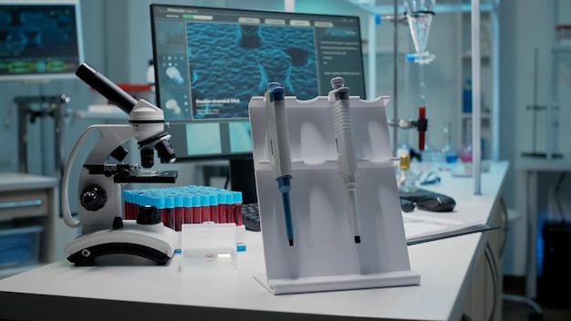 化学実験室での科学的なマイクロピペットと機器のクローズアップ。バキュテナー、試験管、ガラスサンプル、顕微鏡、コンピューター上のDNAアニメーションを備えた空の部屋の医療デスク