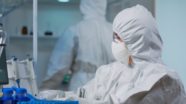 医療研究所で働いているPCでタイピングし、革新的な薬を開発している保護スーツの科学者のクローズアップ。新しいウイルスに対して、科学研究のためのハイテク化学ツールを使用している医師