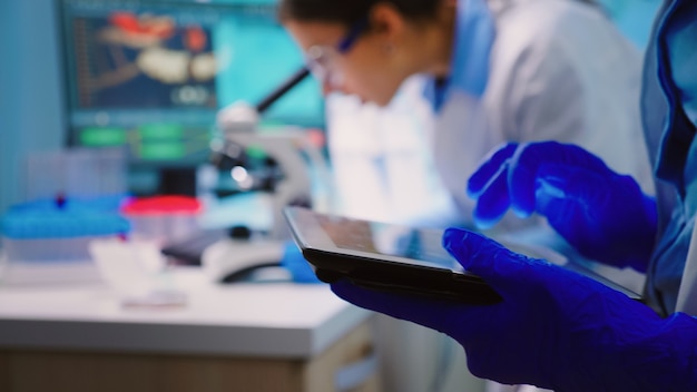 Close up of scientist writing on tablet while team of biologists doing biological researches under microscope in backgorund late at night