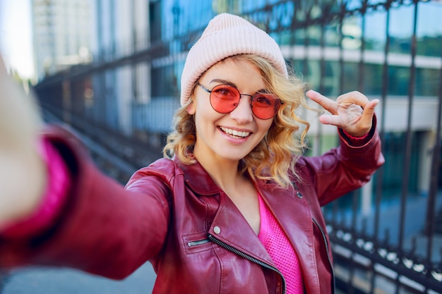 Close up self portrait of joyful hipster enthusiastic woman in trendy pink hat, leather jacket . Showing signs by hand.