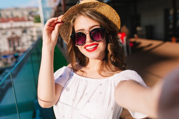 Close-up selfie-portrait of pretty girl with long hair standing on sunlight on terrace. She wears white dress, hat, red lipstick, sunglasses. She is touching hat and smiling.