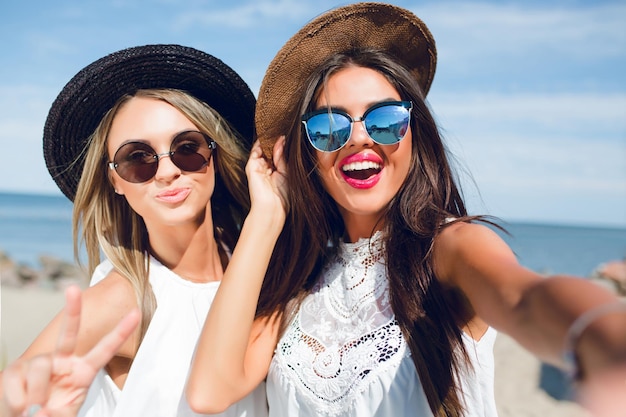 해변에 서 있는 긴 머리를 가진 두 명의 매력적인 갈색 머리와 금발 소녀의 클로즈업 셀카 초상화. 그들은 모자, 선글라스 및 흰색 드레스를 착용합니다. 그들은 카메라에 웃 고 있습니다.