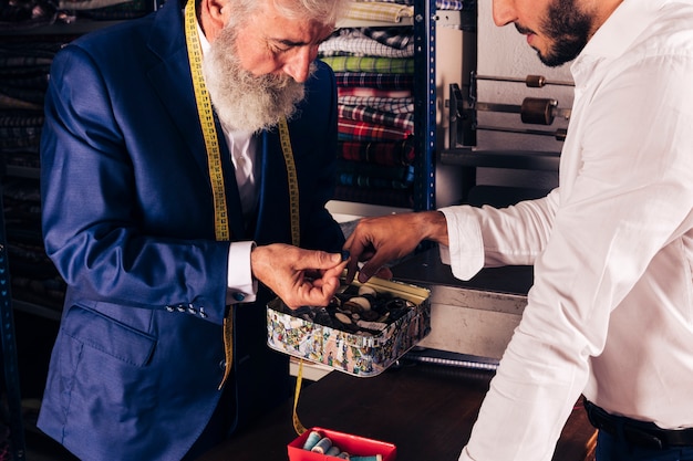 Free photo close-up of a senior male tailor with measuring tape around his neck showing button to his customer