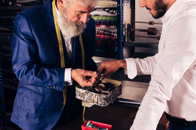 Close-up of a senior male tailor with measuring tape around his neck showing button to his customer