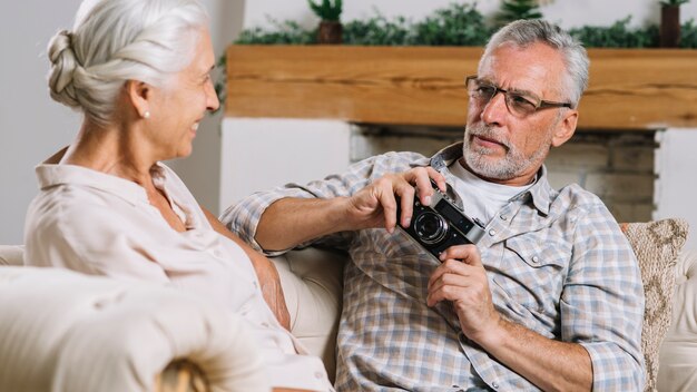 Close-up of senior man holding camera in hand looking at her wife sitting on sofa