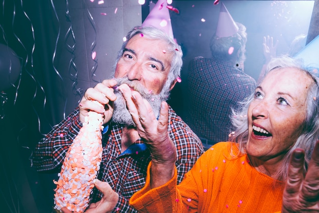 年配の男性がシャンパンのボトルと誕生日パーティーで空気中に紙吹雪を投げる彼女の妻を保持