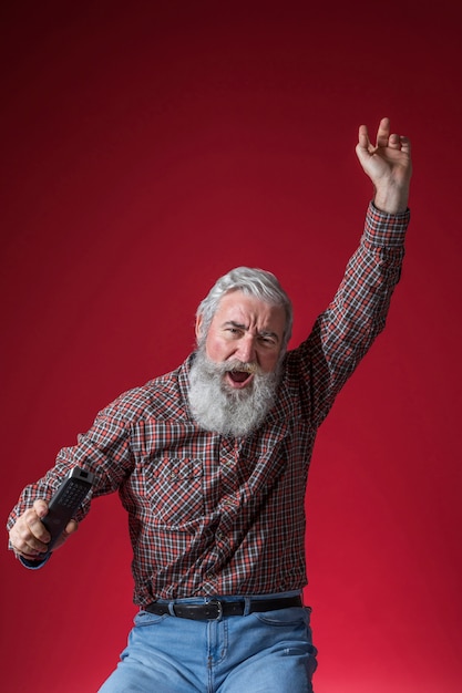 Close-up of a senior man holding remote control in hand cheering against red background