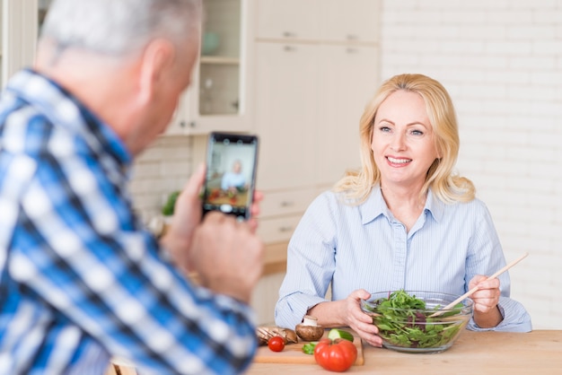 Close-up of a senior man taking photo of her wife preparing fresh salad in glass bowl