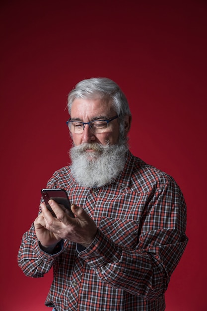 Close-up of a senior man with grey beard using the mobile phone standing against red background