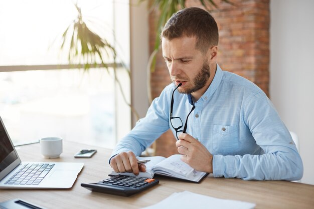 Close up of serious finance manager spending morning in office, holding glasses in mouth, looking at calculation results