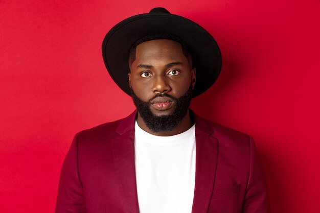 Close-up of serious-looking african american guy in blazer and black hat, looking at camera, standing over red background