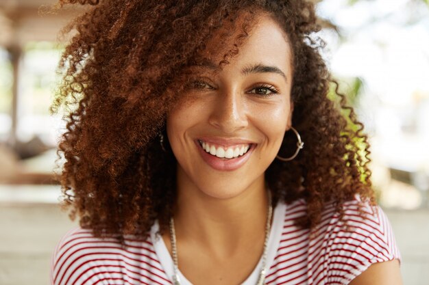 Close up shot of adorable African American woman has broad smile, wears striped t shirt, being in good mood, rests in cafeteria with best friends. Smiling dark skinned young female poses indoor