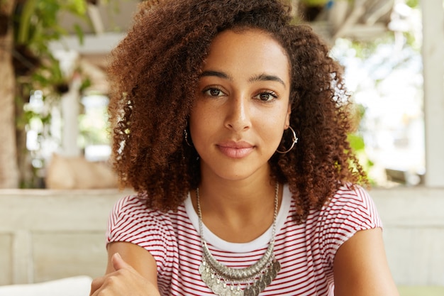 Foto gratuita immagine ravvicinata di adorabili femmine dalla pelle scura con aspetto fiducioso, ha i capelli ricci, medita su qualcosa. la donna afroamericana ha resto contro l'interno del caffè.