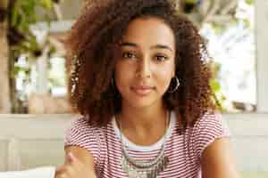 Free photo close up shot of adorable dark skinned females with confident look, has curly hair, ponders about something. african american woman has rest against cafe interior.