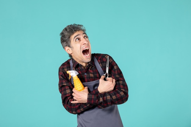 Close up shot of afraid hairdresser wearing gray apron and holding scissors spray bottle on pastel blue color background