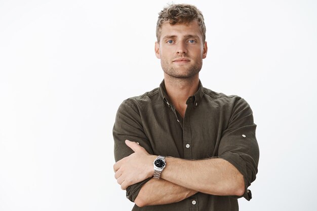 Close-up shot of ambitious, successful and determined handsome rich bachelor with blue eyes and blond stylish haircut in watch and olive shirt cross hands