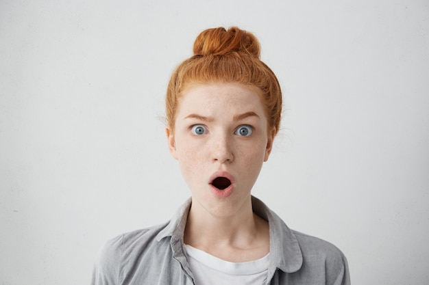 Close up shot of astonished freckled teenage girl wearing her ginger hair in bun raising eyebrows and opening mouth with startled look, absolutely shocked