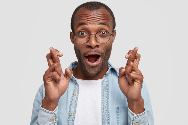 Close up shot of attractive African American amazed male keeps fingers crossed