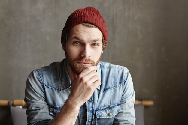 Foto gratuita chiuda sul colpo di giovane maschio barbuto europeo bello attraente vestito in camicia di jeans alla moda e cappello marrone che sorridono, avendo lo sguardo premuroso, profondo e saggio, toccando la sua barba