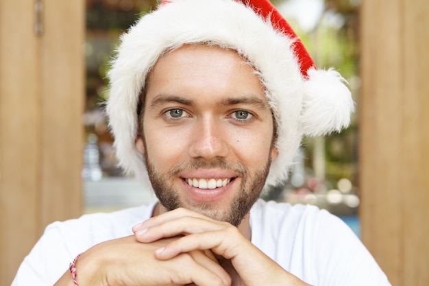 Foto gratuita chiuda sul colpo del giovane attraente con la barba lunga che indossa il cappello di babbo natale che guarda l'obbiettivo e sorride allegramente, aspettando la festa di capodanno mentre si godono le vacanze felici nel paese tropicale