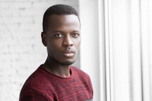 Close up shot of attractive young dark-skinned man in casual sweater standing at window