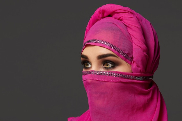 Close-up shot of an attractive young female with an expressive smoky eyes wearing the chic pink hijab decorated with sequins. She has turned her head and looking away on a dark background. Human emoti