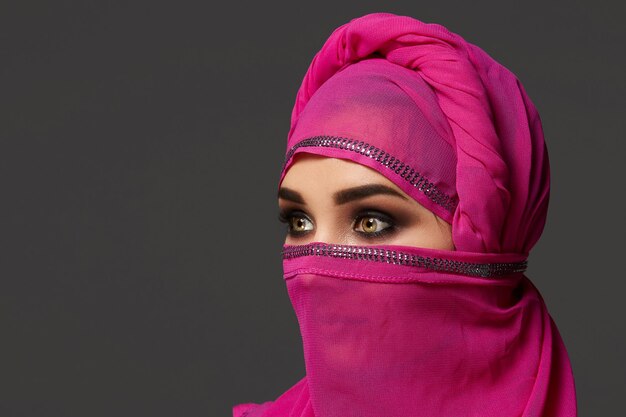 스팽글로 장식된 세련된 분홍색 히잡을 쓰고 표정 연기가 자욱한 눈을 가진 매력적인 젊은 여성의 클로즈업 샷. 그녀는 고개를 돌려 어두운 배경을 바라보고 있습니다. 인간 이모티콘