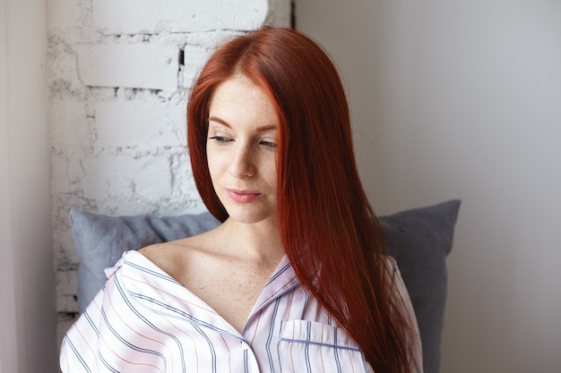 Close up shot of attractive young female with perfect freckled skin and long shiny ginger hair sitting at home with pillow behind her back, dressed in striped nightsuit slipping down her shoulder