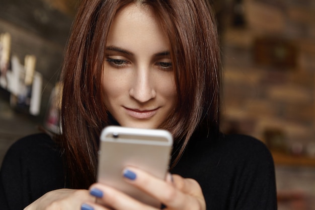 Close up shot of attractive young woman with straight brunette hair looking at screen of her phone