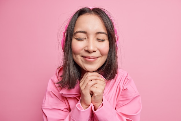 Close up shot of beautiful Asian teenage girl keeps hands under chin closes eyes with pleasure listens via wireless headphones enjoys favorite melody isolated over pink wall. Hobby concept